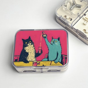 Pode incluir: Um estojo de comprimidos de plástico transparente com uma tampa rosa com uma ilustração de desenho animado de dois gatos em uma mesa. Um é preto e branco, o outro azul, e eles brindam com bebidas. O estojo tem um interior branco.