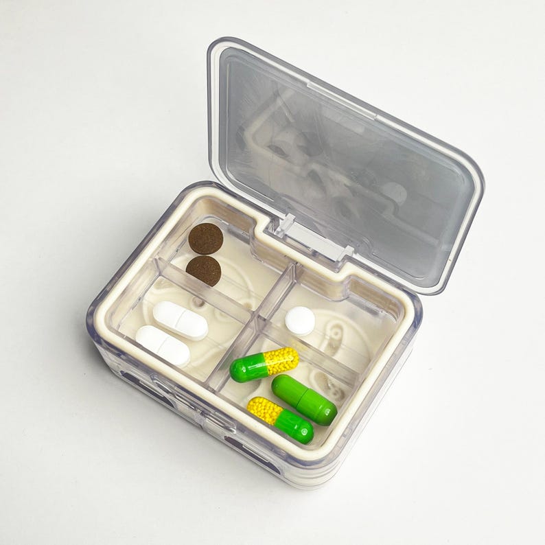 May include: A clear, divided pill organiser with a hinged lid, containing various pills and capsules. The organiser has multiple compartments holding brown, white, and green and yellow capsules. The lid is partially open.