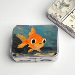 May include: A clear plastic case with a painted image of an orange goldfish. The goldfish has large, expressive eyes and is set against a blue and green background. The case is rectangular and appears to be a storage container.