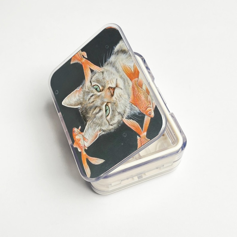 May include: A clear, rectangular pill box with a hinged lid. The lid features a graphic of a grey tabby cat with green eyes surrounded by orange goldfish against a black background. The box is open.