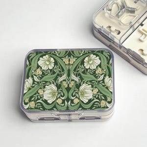 May include: A clear plastic pill organizer with a green floral design on the lid. The design features white flowers and green leaves on a dark green background. Another organizer is partially visible in the background.