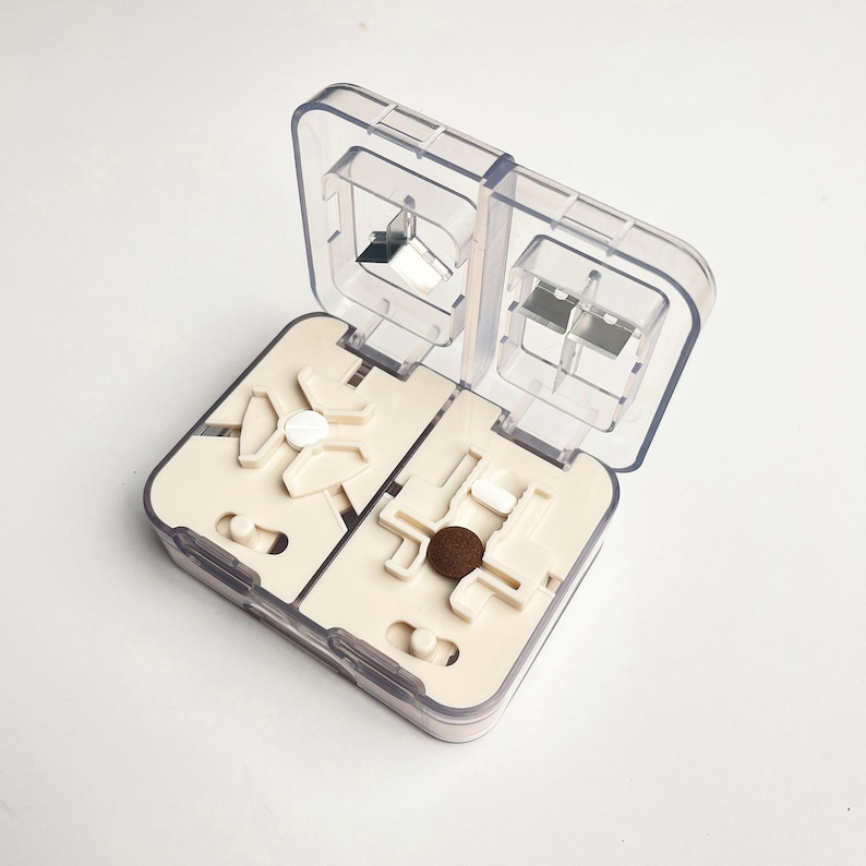 May include: A clear, hinged pill organiser with two compartments. Each compartment has a beige interior with pill-holding slots and a pill cutter. The organiser is open, revealing various pills and a brown tablet.