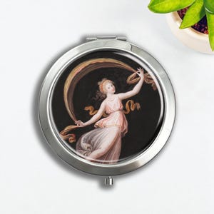 Puede incluir: Un espejo compacto redondo y plateado con una pintura clásica de una mujer con un vestido rosa fluido y una cinta dorada sobre un fondo negro. El compacto está cerrado y tiene un pequeño botón.