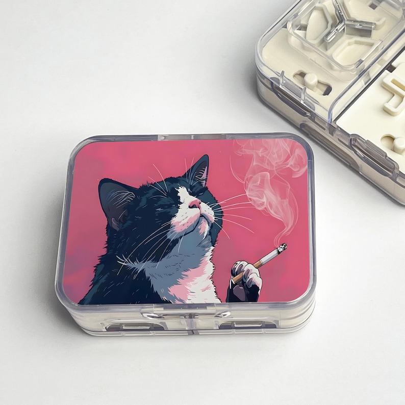 May include: A clear plastic pill case with a pink background featuring a black and white cat smoking a cigarette. The cat is looking up with smoke rising from the cigarette. Another clear plastic case is in the background.