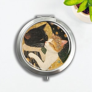 Op de afbeelding: Een zilveren compacte spiegel met een schilderij van twee kussende katten. Eén kat is zwart en de andere is wit en oranje. De achtergrond heeft gouden en donkergroene accenten. De spiegel is rond.