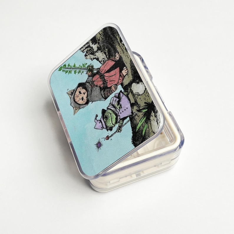 Puede incluir: Una caja de pl&aacute;stico transparente con bisagras y una colorida ilustraci&oacute;n. La tapa muestra dos personajes fant&aacute;sticos, uno parecido a un gato y otro a una rana, sobre un fondo azul claro. La caja est&aacute; abierta, revelando su interior vac&iacute;o.
