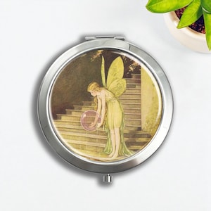 May include: A silver compact mirror featuring a vintage illustration of a fairy in a flowing green dress with yellow wings, holding a pink orb. The background shows a staircase and soft, muted colors.