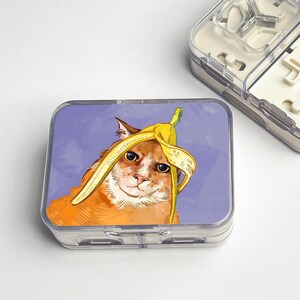 May include: A clear plastic pill case with a humorous illustration of a ginger cat wearing a banana peel hat. The cat is set against a lavender background. Another clear case is partially visible.
