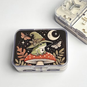 May include: A rectangular, clear plastic pill case with a fantasy design. The design features a green frog in a witch's hat on a red and white mushroom, with a crescent moon and butterflies on a black background. The case is rectangular.