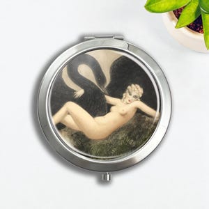 Puede incluir: Un espejo compacto redondo y plateado con una ilustración vintage de una mujer desnuda y un cisne negro. La obra de arte tiene un estilo clásico, con colores apagados y un diseño detallado. Un accesorio práctico.