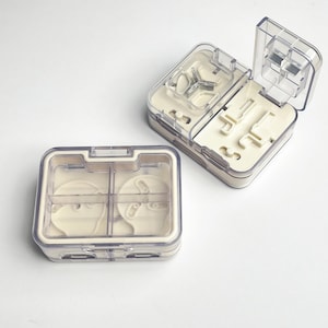 May include: Two clear plastic pill organisers with off-white interiors. One is open, revealing compartments with various shapes. The other is closed, showing a divided interior. Both have a rectangular shape and are designed for medication storage.
