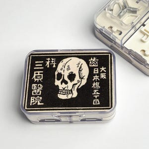 Puede incluir: Un contenedor de plástico transparente con un diseño de calavera en blanco y negro y texto japonés. El contenedor rectangular está sobre una superficie blanca y probablemente contiene objetos pequeños.
