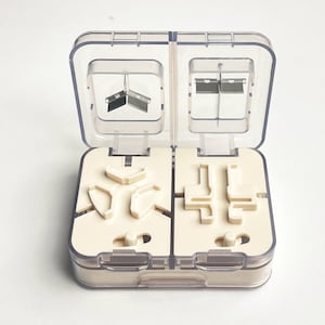 May include: A clear plastic pill cutter with a hinged lid, open to reveal a cream-coloured interior with various compartments and blades. The blades are silver and the compartments are designed to hold pills for cutting.