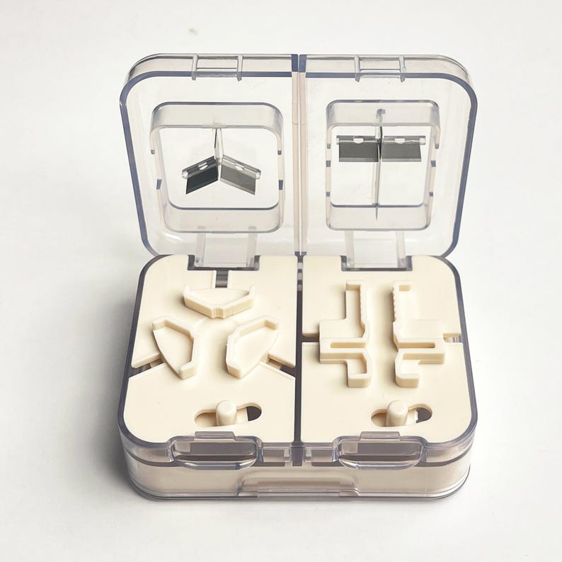 May include: A clear plastic pill cutter with a hinged lid, open to reveal a cream-coloured interior with various compartments and blades. The blades are silver and the compartments are designed to hold pills for cutting.