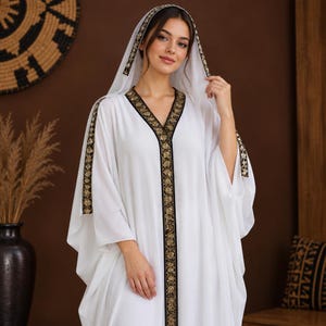 Könnte beinhalten: Ein weißer Kaftan mit V-Ausschnitt und angesetzter Kapuze, verziert mit einer schwarz-goldenen Stickborte entlang der vorderen Knopfleiste, den Kapuzenrändern und den Ärmelbündchen. Das Kleidungsstück hat lockere, fließende Ärmel und eine lange Silhouette.