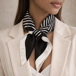 Puede incluir: Un pañuelo de seda a rayas blancas y negras anudado al cuello. El pañuelo presenta un llamativo patrón de rayas negras sobre fondo blanco, con un borde blanco. Se lleva con una blazer color crema.