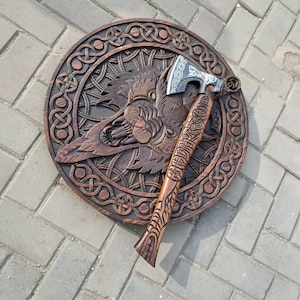 May include: A wooden, circular wall hanging with a carved wolf design and intricate knotwork border. An axe with a detailed wooden handle and silver blade rests on the wall hanging. The wood has a warm, brown tone.