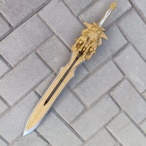 May include: A decorative sword with a gold-colored hilt and blade. The hilt features intricate carvings and a detailed design. The blade has a silver finish with gold accents. The sword is displayed on a gray brick surface.