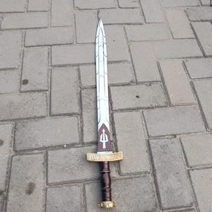 May include: A decorative sword with a white blade featuring a geometric design. The hilt is brown with gold accents and a gold crossguard. The text "ANAKLUMOS" is visible on the crossguard. The sword is lying on a gray brick surface.