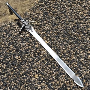 May include: A long, silver sword with a black handle and ornate silver crossguard featuring a skull design. The blade is straight and sharp, with a pointed tip. The sword is displayed on a textured, pebble-like surface.