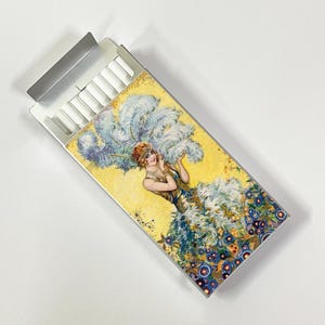 May include: A cigarette box with a vintage illustration of a woman in a feathered dress. The box has a yellow background with blue and gold floral accents. The cigarettes are visible inside the open box.