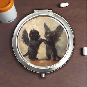 Op de afbeelding: Een zilveren compacte spiegel met een grillige illustratie van twee zwarte kittens met vleermuisvleugels, die elkaars hand vasthouden. Het kunstwerk heeft een vintage, schilderachtige stijl met een warme, neutrale achtergrond. De compact is rond en heeft een scharnierende opening.