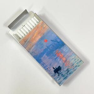 May include: A metal cigarette case with a hinged lid, partially open to reveal white cigarettes. The case is decorated with a painting of a sunset over water, featuring blue, orange, and red hues.