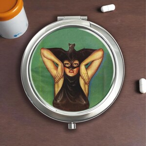 Peut inclure: Un miroir de poche rond et argenté avec un couvercle à charnière. L'image du miroir représente une femme dans un costume de chauve-souris noir sur un fond vert. La femme a les bras levés. Le compact est sur une surface brune.