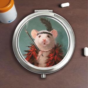 Puede incluir: Un espejo compacto plateado con una ilustración caprichosa de un ratón blanco vestido con un atuendo de flapper, con una pluma, un collar de perlas y un boquilla. El ratón está sobre un fondo turquesa.