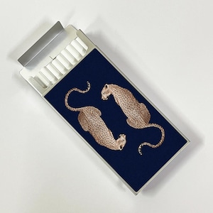 May include: A rectangular silver metal cigarette case featuring a dark navy blue panel on the front. The panel displays two illustrated leopards with tan fur and black spots, positioned facing each other. The case has a hinged lid and internal slots.