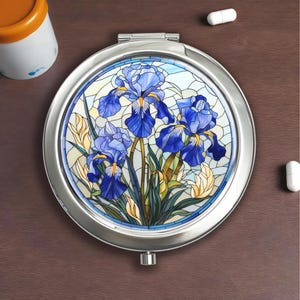 May include: A round, silver compact mirror with a stained-glass design featuring blue iris flowers. The flowers have yellow accents and are set against a light blue and white background. The mirror is open, revealing the design.