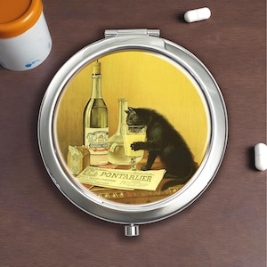 May include: A round, silver compact mirror with a vintage-style image of a black cat drinking from a glass. The image includes a bottle, a newspaper with the text "PONTARLIER", and a yellow background.
