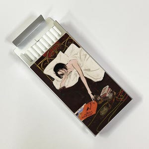 May include: A cigarette pack with a silver lid, partially open, revealing white cigarettes. The pack's design features an illustration of a person lying in bed, reaching for items on a nightstand. The nightstand has a record album and other items.