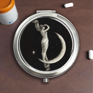 Puede incluir: Un espejo compacto plateado con un fondo negro que representa a una mujer sobre una luna creciente. La mujer lleva un vestido fluido, con estrellas y elementos celestiales. El espejo redondo tiene una apertura con bisagras.