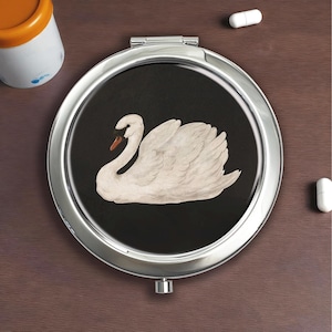 Op de afbeelding: Een zilveren compacte spiegel met een zwarte achtergrond met een witte zwaan illustratie. De zwaan heeft een gebogen nek en oranje snavel. De compact is rond en heeft een scharnierend deksel.