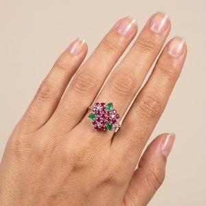 Peut inclure: Une bague ornée d'un groupe de rubis rose foncé taille ronde et de trois émeraudes vertes taille poire disposées en motif floral, sur un anneau métallique fin. Les lettres NNJ sont imprimées en petit texte blanc près de la base de la monture.