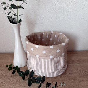 Könnte beinhalten: Ein beiger Stoffkorb mit weißem Tupfenmuster oben und vertikal gestreiftem Boden. Eine weiße Vase mit grünen Blättern und ein Zweig Eukalyptus stehen neben dem Korb auf einer Holzoberfläche.