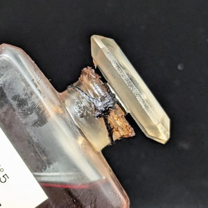 Pu&ograve; includere: Una bottiglia di vetro trasparente con un liquido rosso scuro, accanto a un cristallo giallo chiaro. La bottiglia ha un bordo rotto con materiale scuro e carbonizzato. Il cristallo &egrave; allungato e sfaccettato, con un aspetto leggermente torbido.