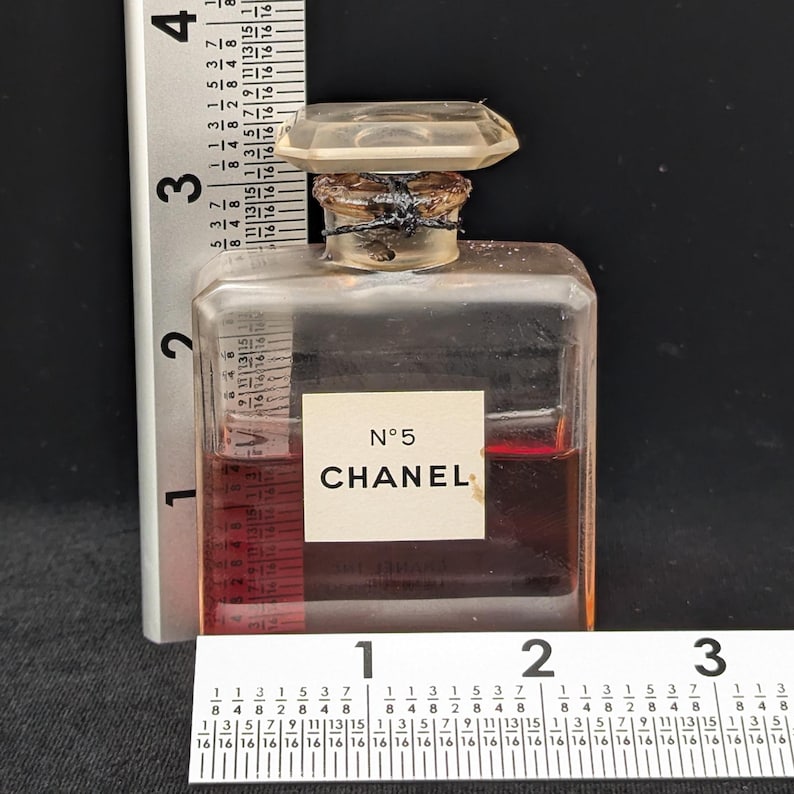 Pu&ograve; includere: Un flacone di profumo Chanel No. 5 vintage. Il flacone rettangolare trasparente ha un tappo trasparente sfaccettato. L'etichetta recita "N&deg;5 CHANEL". Il flacone contiene un liquido marrone rossiccio. Un righello &egrave; posto accanto al flacone.