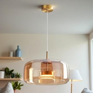 May include: A modern pendant light fixture with a clear, amber-colored glass shade and a gold-toned metal canopy. The light hangs from a white cord and features a cylindrical gold-toned center. The fixture is designed for indoor use and provides ambient lighting.