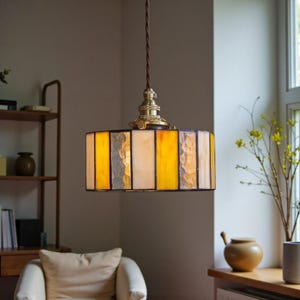 May include: A pendant light fixture with a rectangular shade composed of vertical panels in shades of yellow, cream, and silver. The fixture hangs from a brown cord and has a brass-colored metal base. The light is suspended in a room with a window.