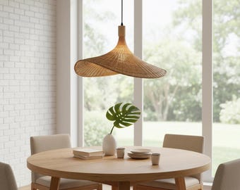 May include: A woven rattan pendant light hangs above a round wooden dining table. The light fixture has a wide, hat-like shade. The table is set with plates, cups, and a vase with a large green leaf. Beige chairs surround the table, and a window is in the background.