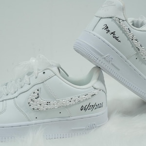 May include: A pair of white sneakers with pearl embellishments on the Nike swoosh and along the heel. The date "06/27/2026" and the name "Mrs. Aadar" are written in black ink on the shoes. The sneakers have white laces and a white sole.