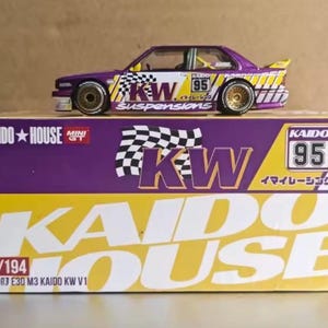 Könnte beinhalten: Ein lilafarbenes und gelbes Miniatur-Rennauto mit dem Text "KW Suspensions" und der Nummer "95" an der Seite, das auf einer Schachtel steht. Die Schachtel ist lila und gelb mit dem Text "KAIDO HOUSE" und "KW".