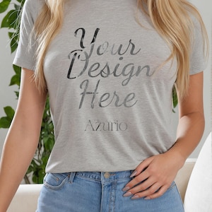 Puede incluir: Una camiseta gris claro con el texto "Your Design Here" en una fuente cursiva. La palabra "Azurio" está debajo del texto. La modelo lleva unos pantalones cortos vaqueros azules.