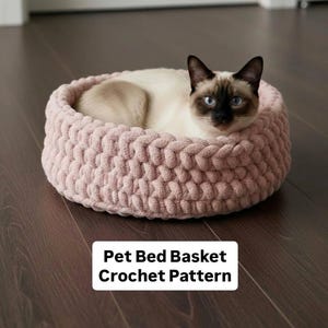 以下が含まれることがあります： ピンク色の太い編み目のペットベッドで休むシャム猫。 丸いバスケットは太い糸でできており、濃い色の木の床に置かれています。 画像のテキストは「Pet Bed Basket Crochet Pattern」と書かれています。