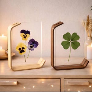 Op de afbeelding: Twee decoratieve lijsten met geperste bloemen. Eén lijst toont gele en paarse viooltjes, de andere een klavertjevier. De lijsten hebben een houten basis en bovenkant, met een helder glazen paneel.