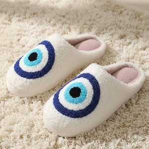 Puede incluir: Un par de zapatillas blancas y esponjosas con un gran diseño bordado de ojo turco en azul, celeste y negro. Las zapatillas tienen un interior suave y rosa, diseñadas para uso en interiores.
