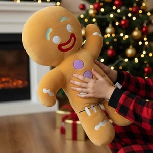 May include: A plush gingerbread man doll with a smiling face, white icing details, and purple buttons. The soft toy is light brown and is being held in front of a Christmas tree.