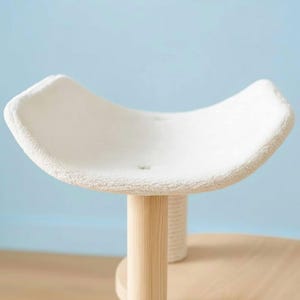 May include: A cat perch featuring a curved, white, plush-covered platform supported by a light-colored wooden post. The platform has a soft, textured surface with two visible tufted indentations, providing a raised, cradle-like shape for resting.